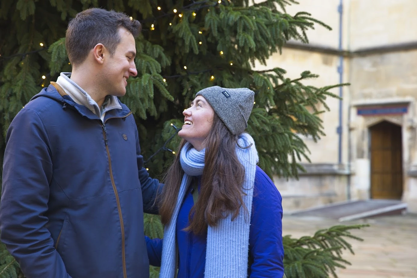 Morgan and Tom smiling at each other in front of a fir tree with fairy lights